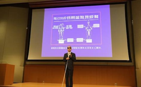セミナー会場の壇上で視覚に障害のある社長が、AIの導入について説明をしている画像です。