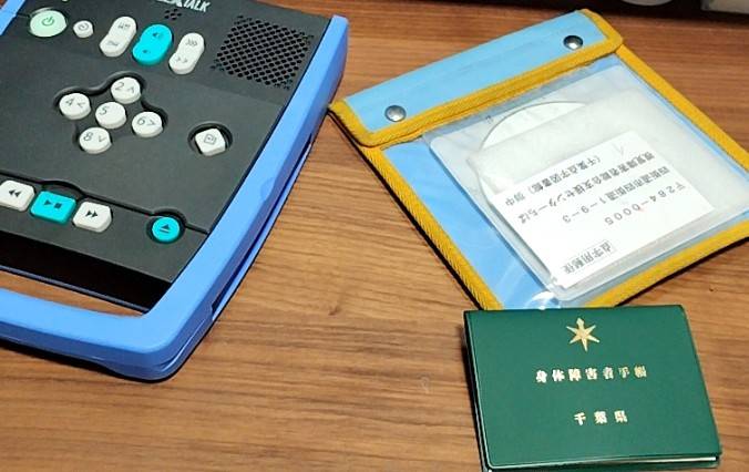 テーブルの上に置かれた左側の機械が視覚障害者が使用するデイジー図書再生機、右側の袋に入っている物が点字図書館からお借りしたデイジー図書専用のCDです。その下においてあるのが緑のケースに入っている身体障害者手帳です。