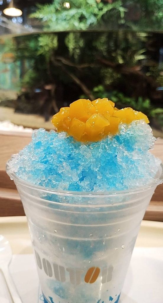 熱帯魚の水槽を背に、「マンゴーブルーハワイかき氷」をメインに撮影した画像で、背景の水槽には様々な小さな熱帯魚が活発に泳いでいます。
