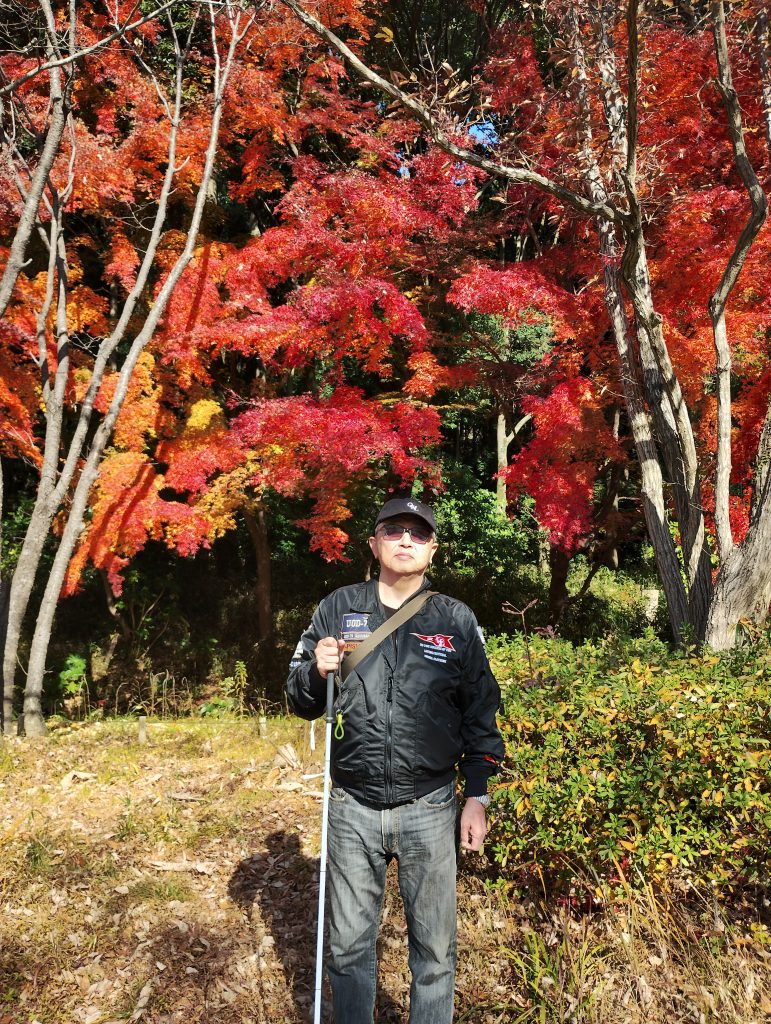 視覚障害者の男性が白杖を右手に持ち、松戸市の「21世紀の森と広場」の紅葉の前で撮影した写真です。