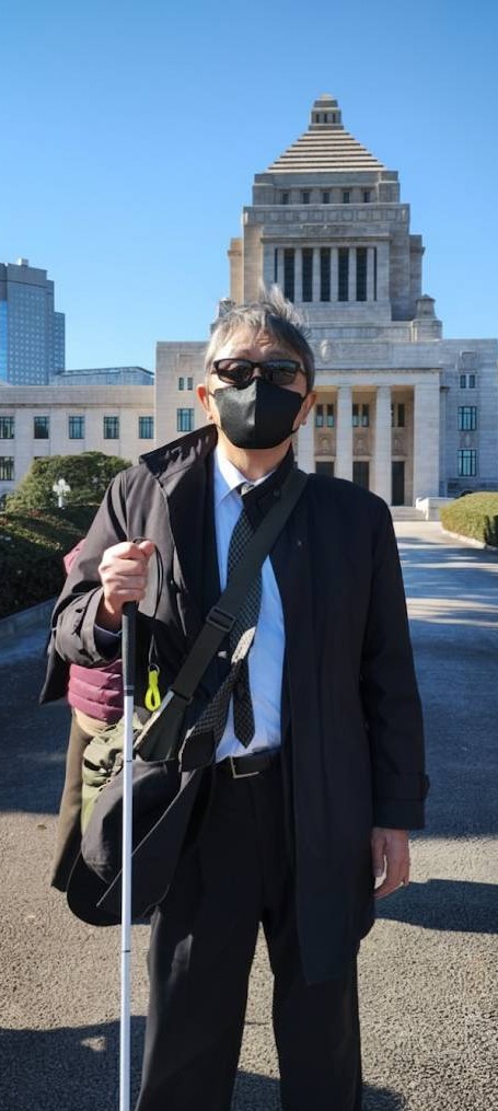視覚障害者であるアイズルームの代表が、白杖を右手に持ち国会議事堂の前で撮影した写真です。