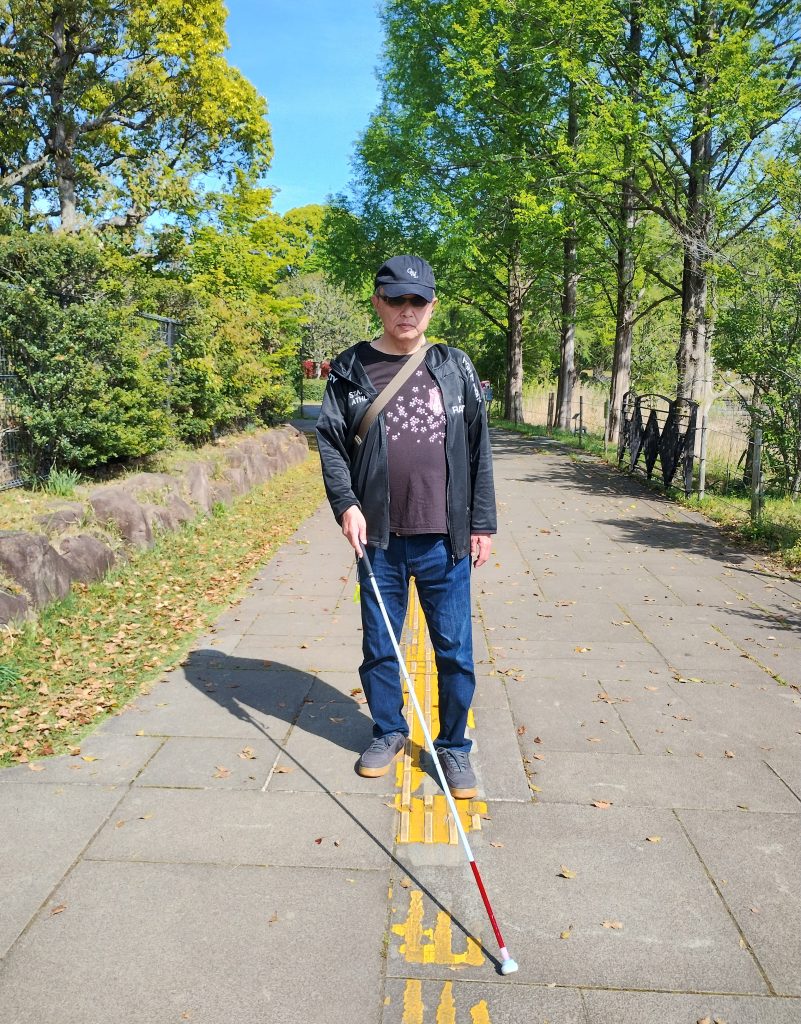春の日差しの強い柏の葉公園を、男性視覚障害者が白杖をつきながら、点字ブロックの上を一人で歩いている画像です。