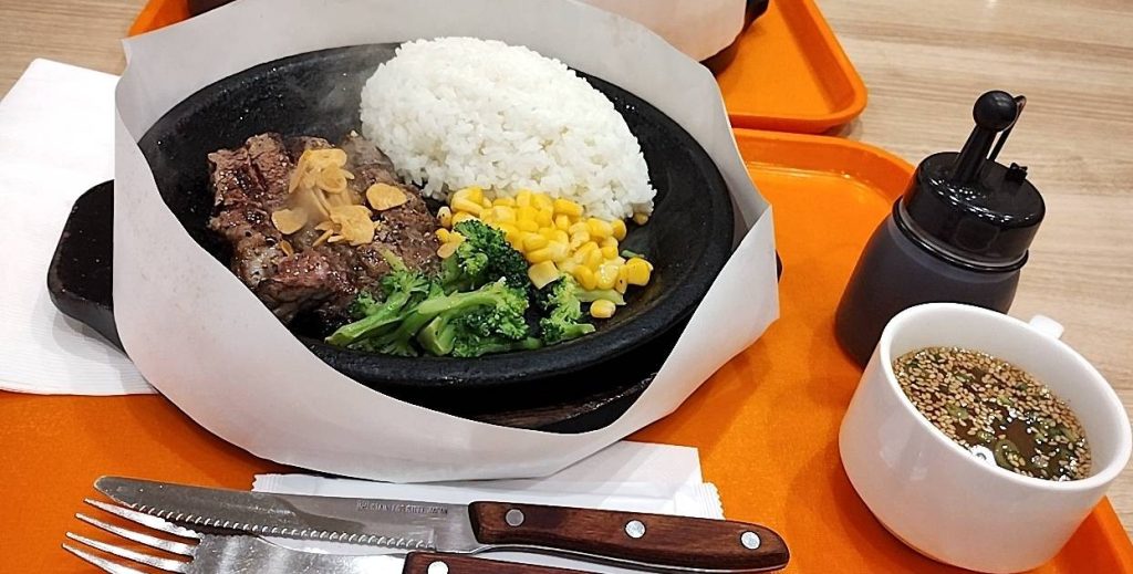 いきなりステーキの格安ランチプレート(スープ付き)お肉の上にニンニクスライスが乗っており、付け合わせはブロッコリーとコーン、大盛りライスの商品画像です。