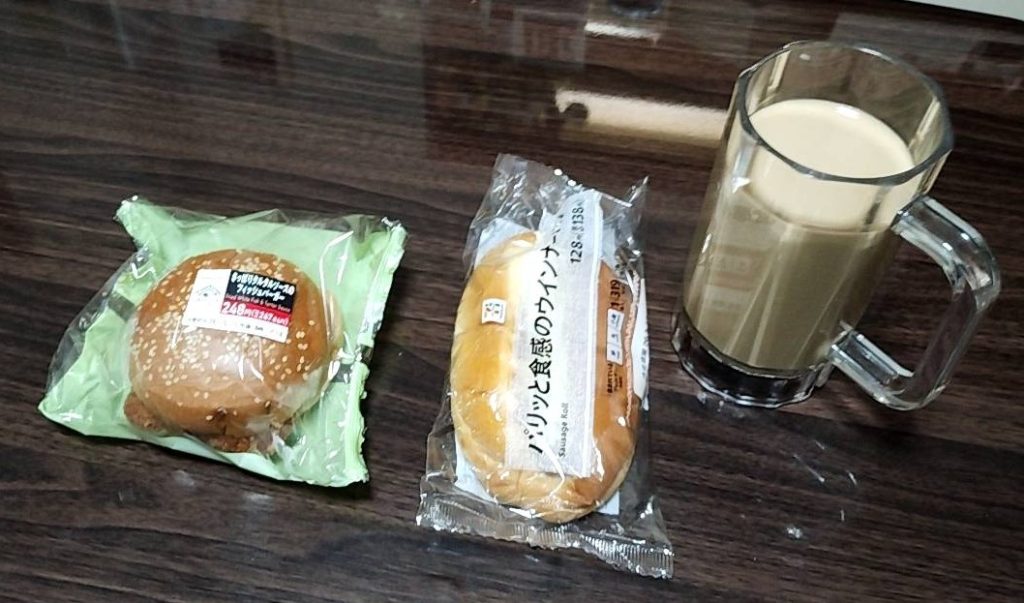セブンイレブンで購入したソーセージとハンバーグの総菜パン、右上にはビールジョッキの中に牛乳を注ぎコーヒーを加えたコーヒー牛乳の画像