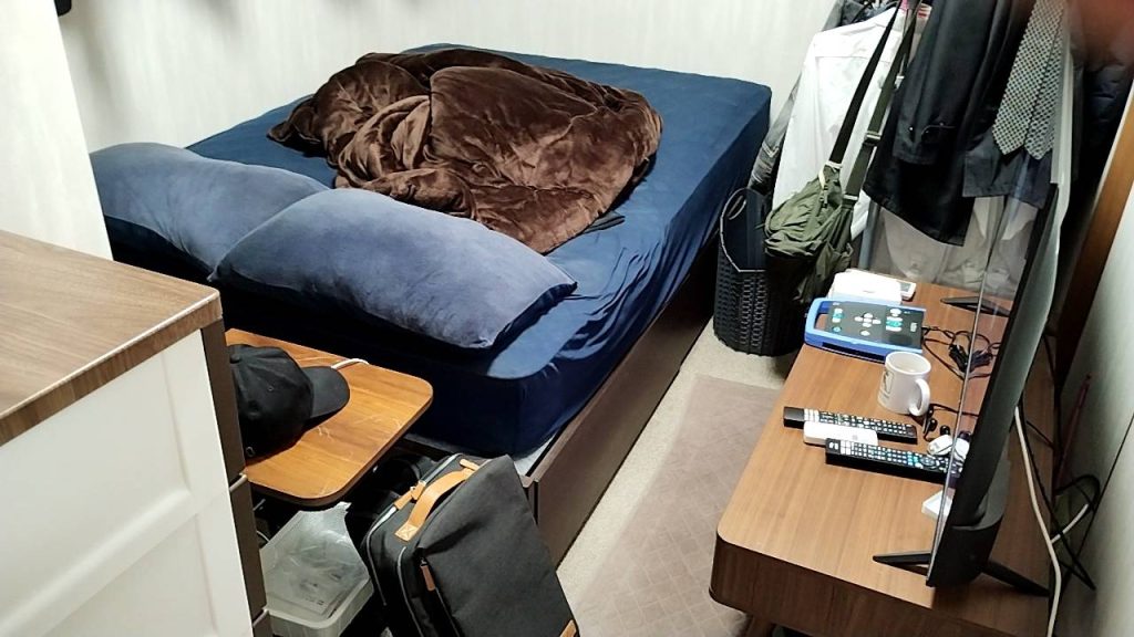 マンションの納戸(窓のない部屋3.5畳)にシモンズベッドとテレビや洋服が、所狭しと置かれている画像です。