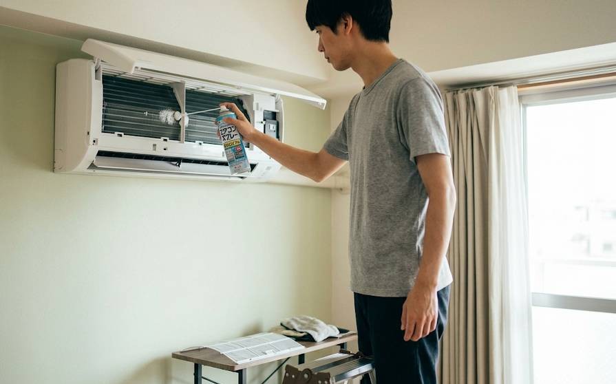 エアコンの除菌・洗浄スプレーを使い、男性が台に乗り家庭用エアコンに向かってスプレーを噴射している画像です。