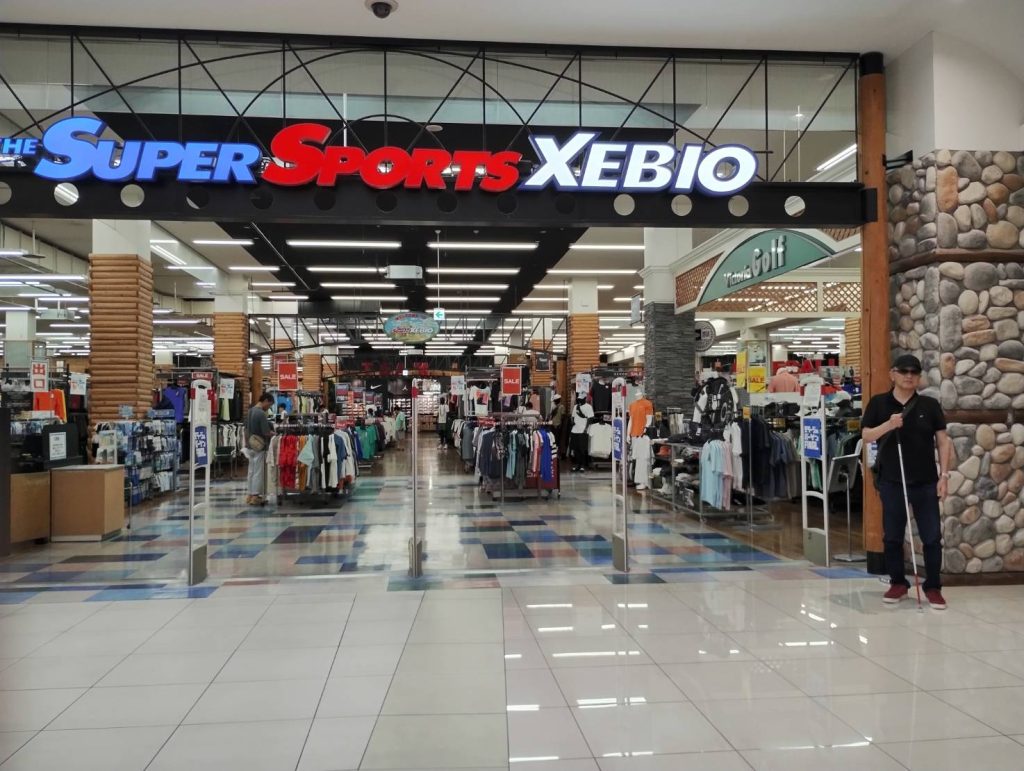 モラージュ柏「スポーツゼビオ」店舗入り口で、黒の刺しゅう入りキャップを被った男性視覚障害者(右手に白杖所持)が写っている写真です。