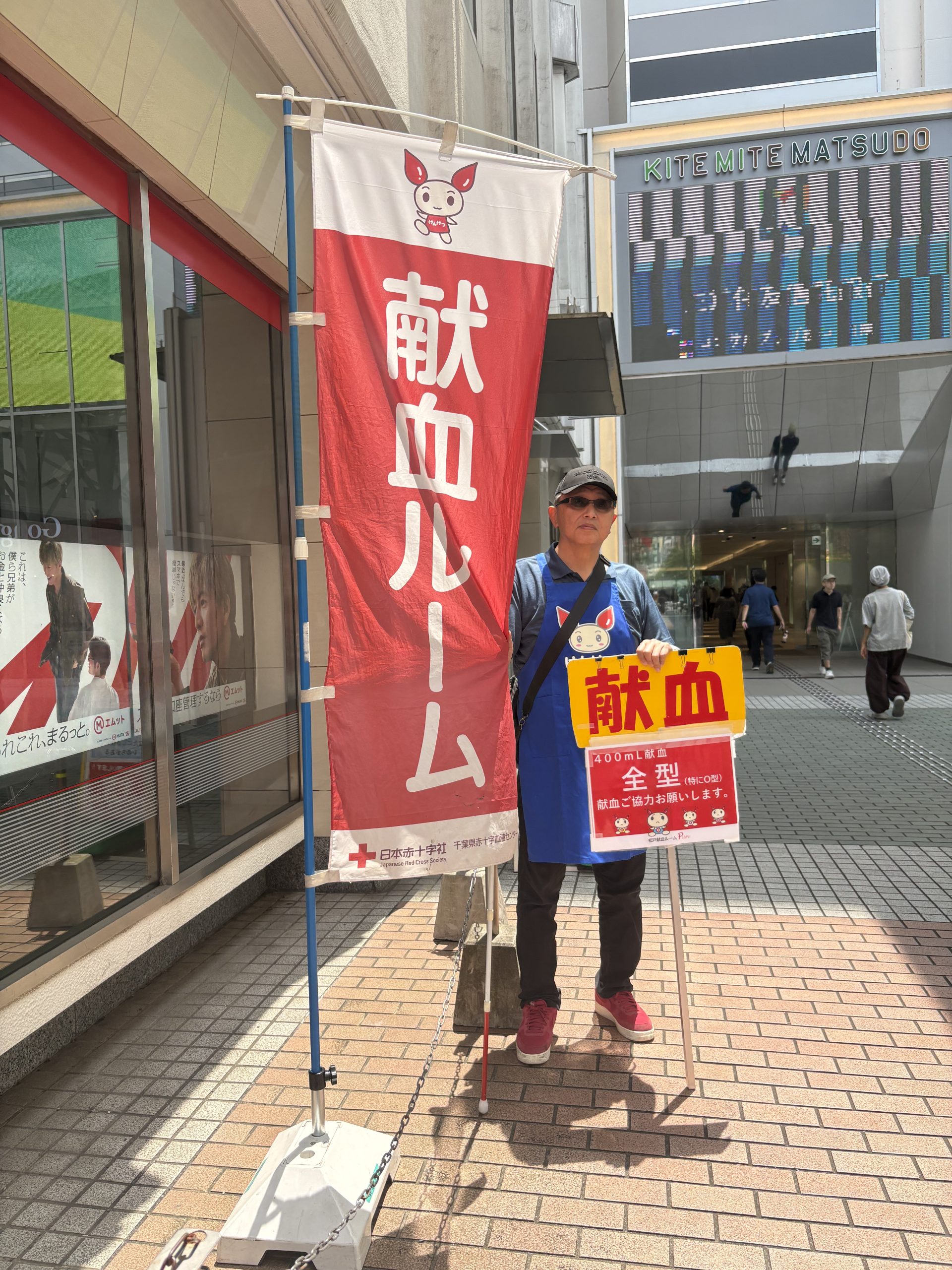 日本赤十字社「松戸献血ルーム」キテミテマツド広場UFJ銀行横の場所で視覚障害者の男性が、献血促進声掛け活動をしている写真です。