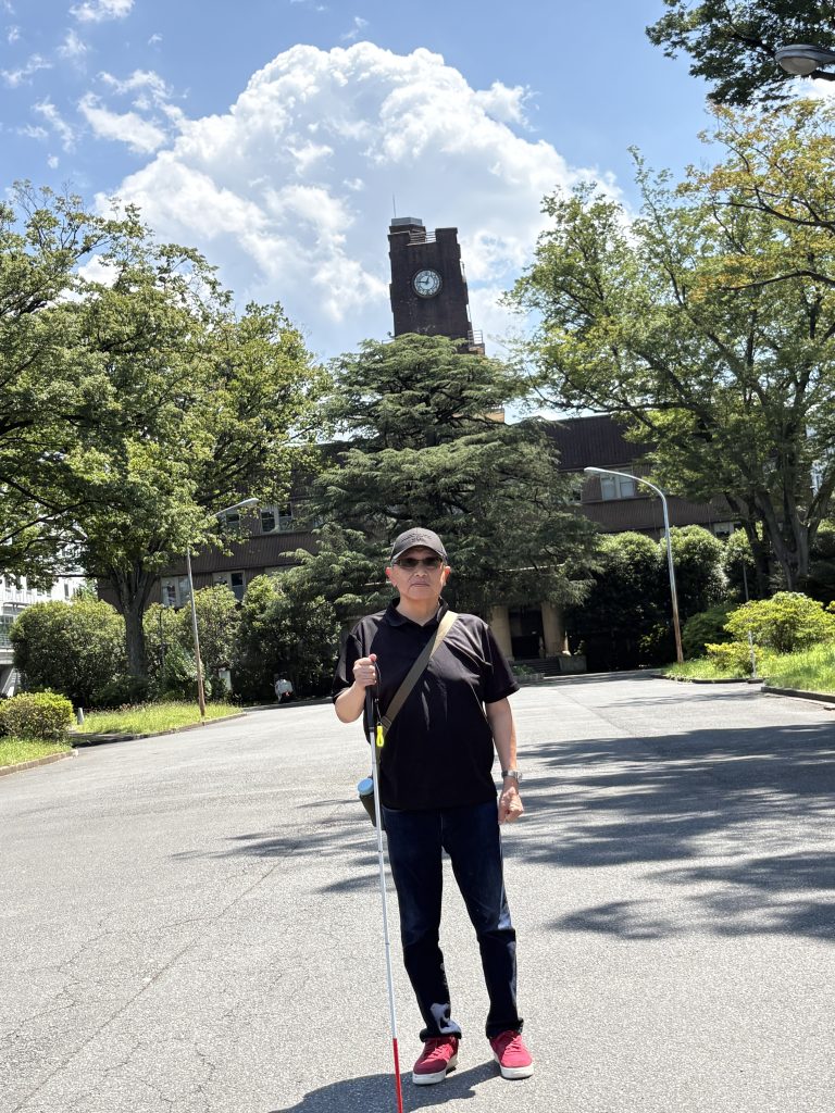 東京大学駒場リサーチキャンパス先端科学技術研究センター時計台前で、視覚障害者の男性が白杖と遮光サングラスを掛けて立っている写真です。