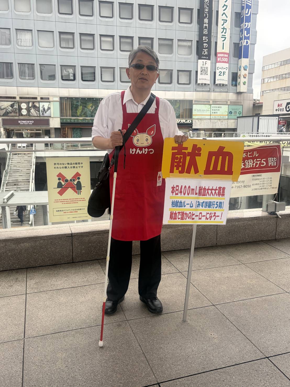福祉支援団体EYESROOM代表の石原幸一が、柏駅前のダブルデッキにて柏献血ルームの献血推進呼び掛け活動ボランティアをしている写真です。視覚障害者の男性が白杖を持ち、献血看板を左手で支えながら赤十字社の赤いエプロンを着けて、写真に写っています。