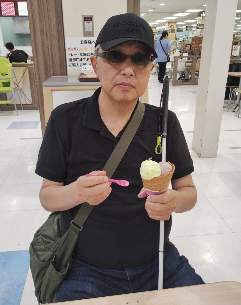 サーティーワンアイスクリームの店内で、視覚障害者の男性がワッフルコーンにトリプルフレーバーをのせたアイスクリームを持っている画像です。