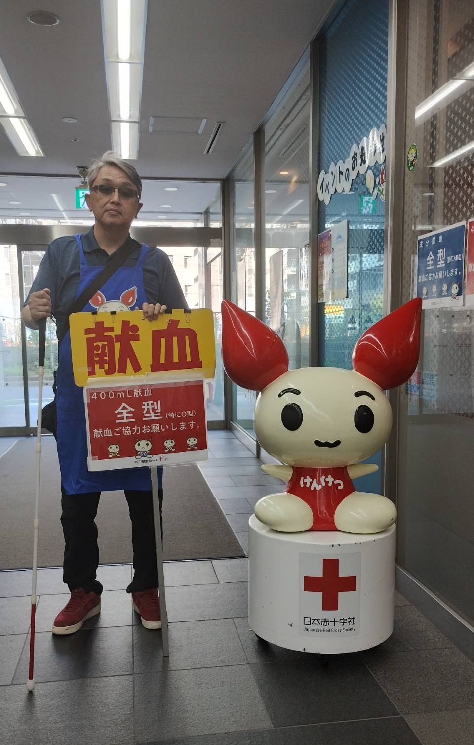 千葉県松戸市「松戸献血ルーム」入り口にある赤十字の献血キャラクターと、私(松戸市視覚障害者協会 副会長石原幸一)と並んで撮影した写真で、献血促進の立て看板も持っています。