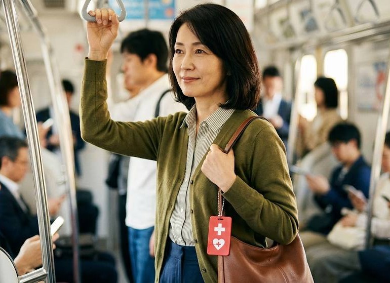 電車内で内部障害を抱えている女性が、ショルダーバックにヘルプマークを付けて立っている画像です。