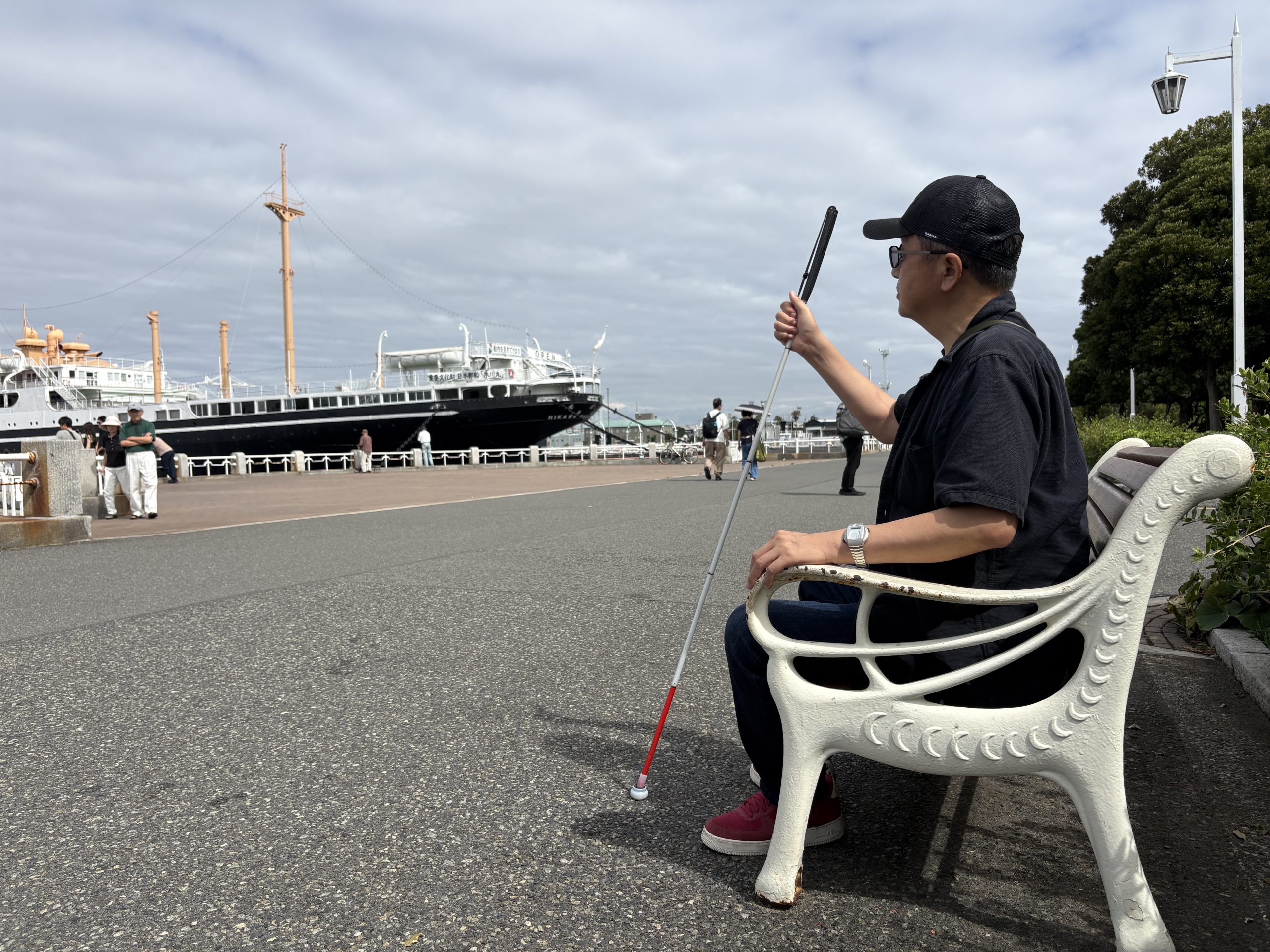 横浜山下公園海沿いの遊歩道に設置してある白いペンキで塗られた鋳物のベンチに視覚障害者の男性が白杖を持って氷川丸を見つめながら座っている画像です。
