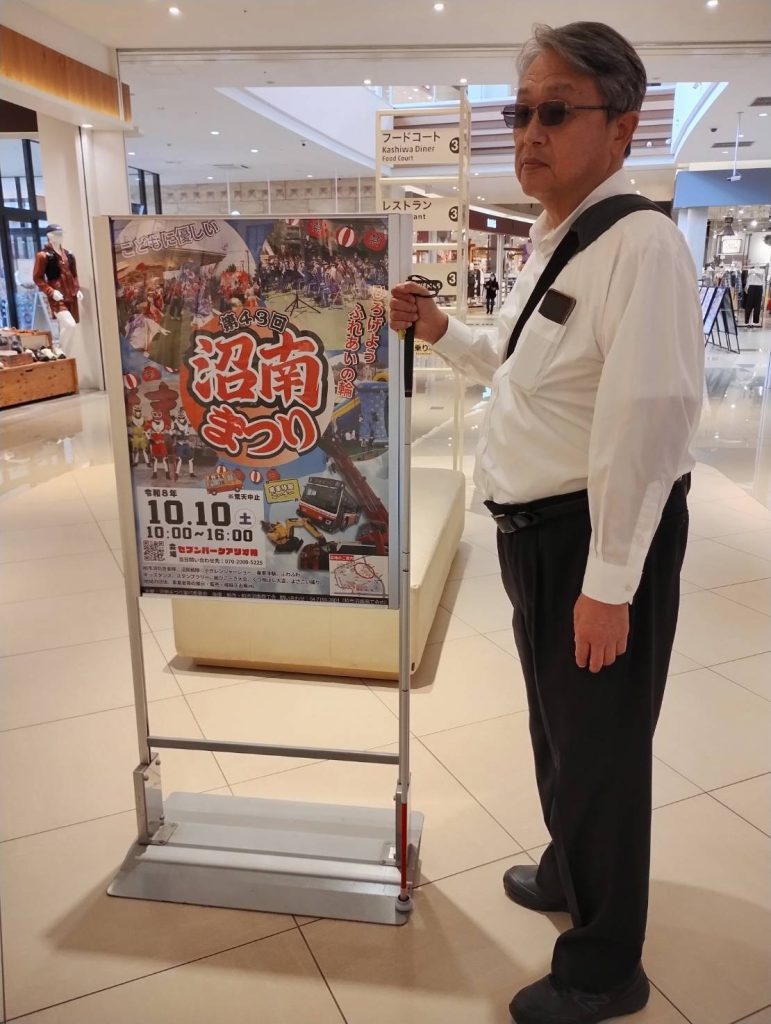 セブンパークアリオ柏「沼南まつり」イベント告知ポスターの横で、視覚障害者の男性が白杖を握りしめ立っている画像です。