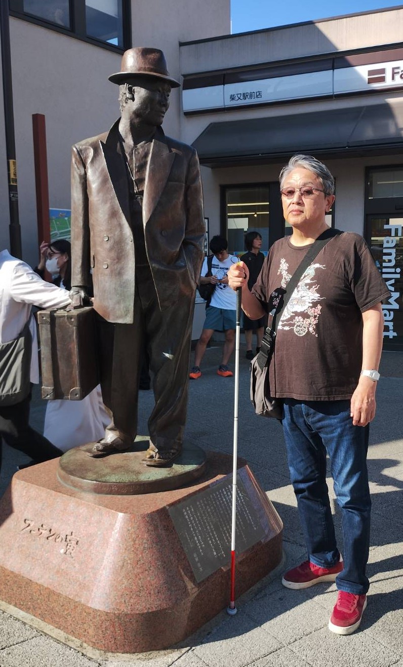 柴又駅前の寅さんの銅像と、男性視覚障害者が写っている写真です。